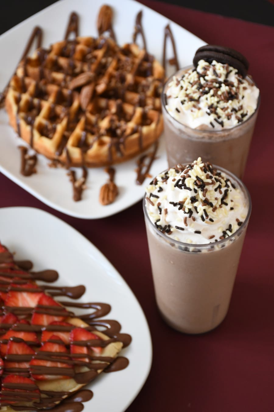 Dos frappés con crema batida junto a un waffle y crepas de fresa